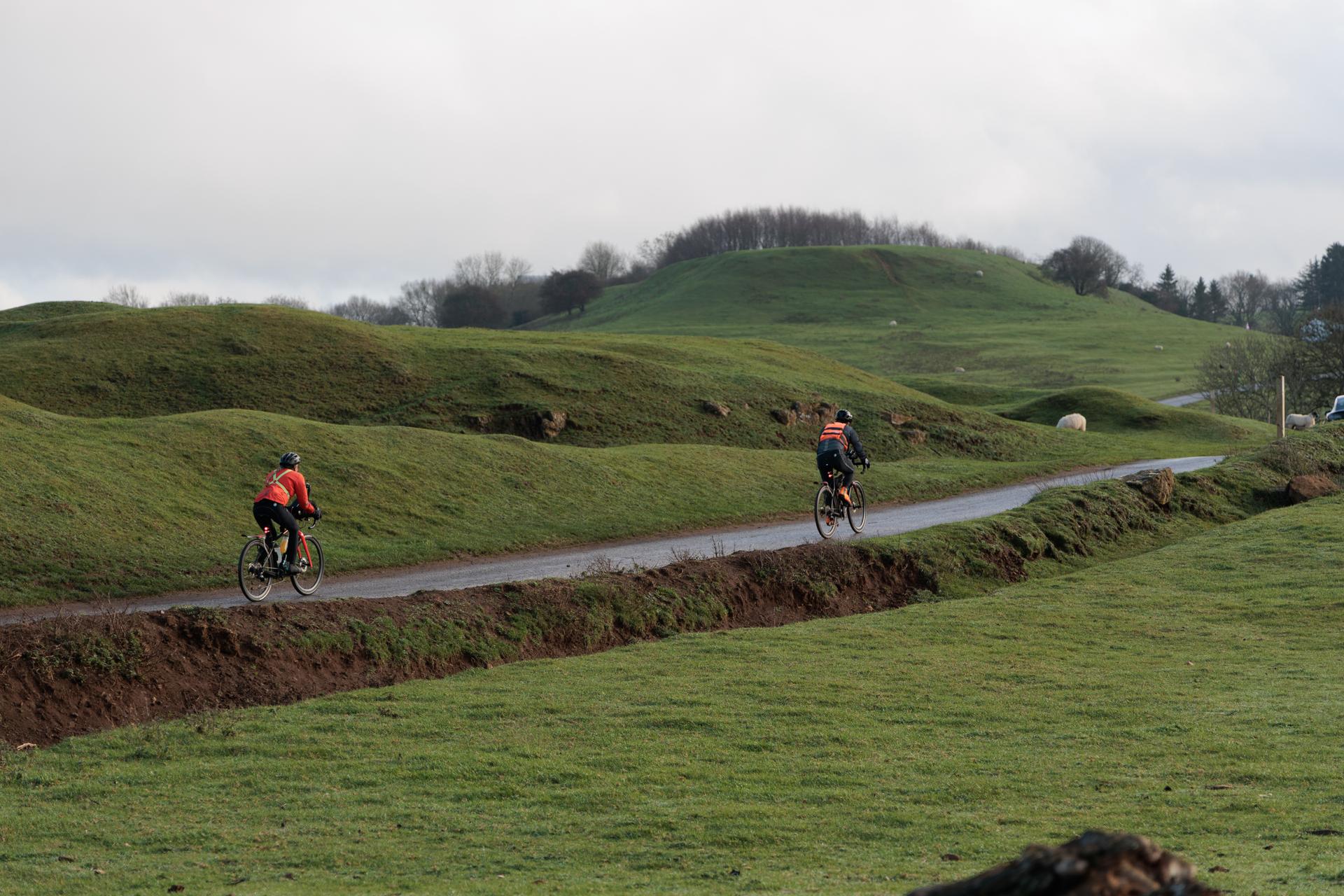 Two cyclists following a narrow lane through uneven green pasture beneath a pale sky.