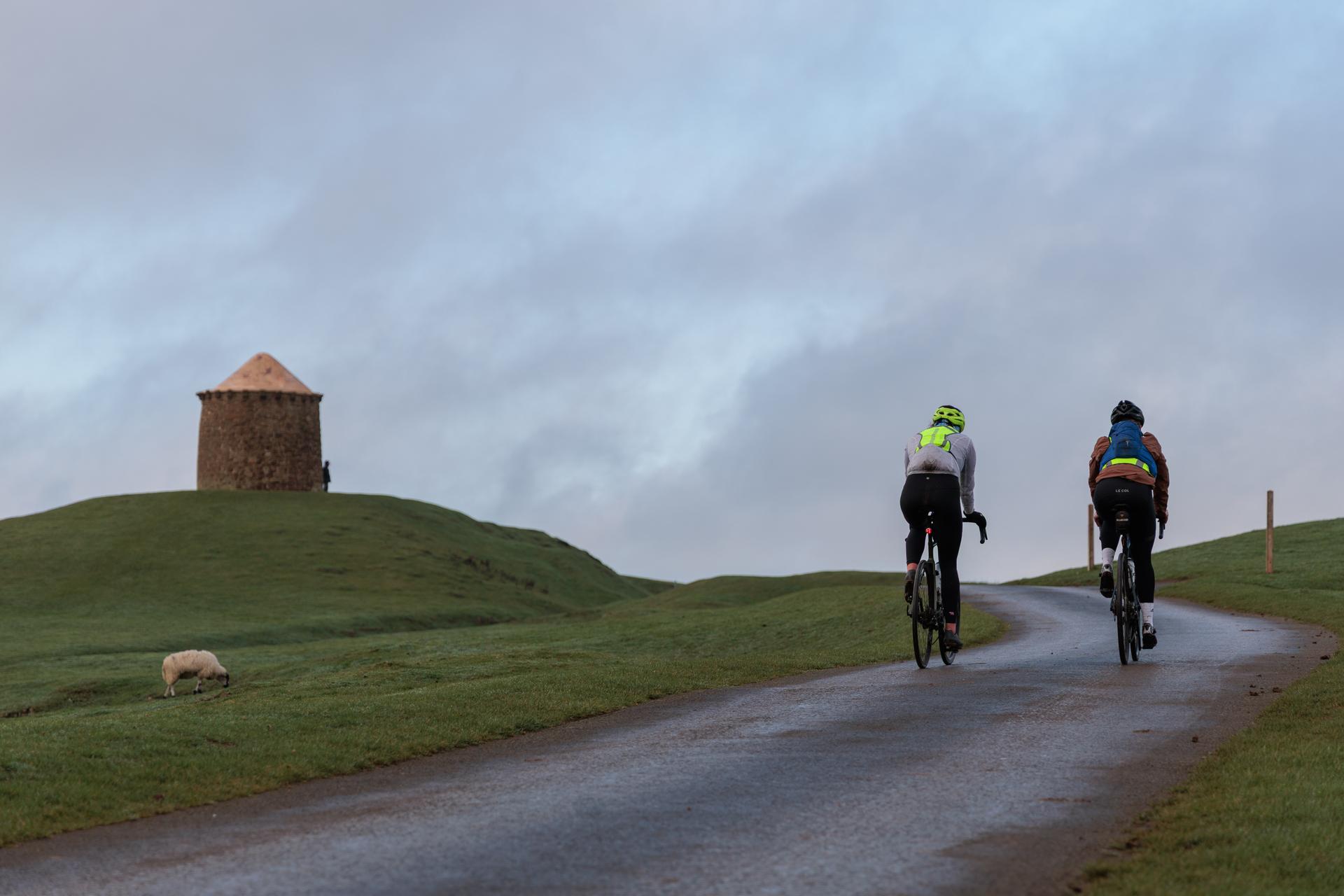 Two cyclists climbing a wet lane beside a stone tower under a grey winter sky.